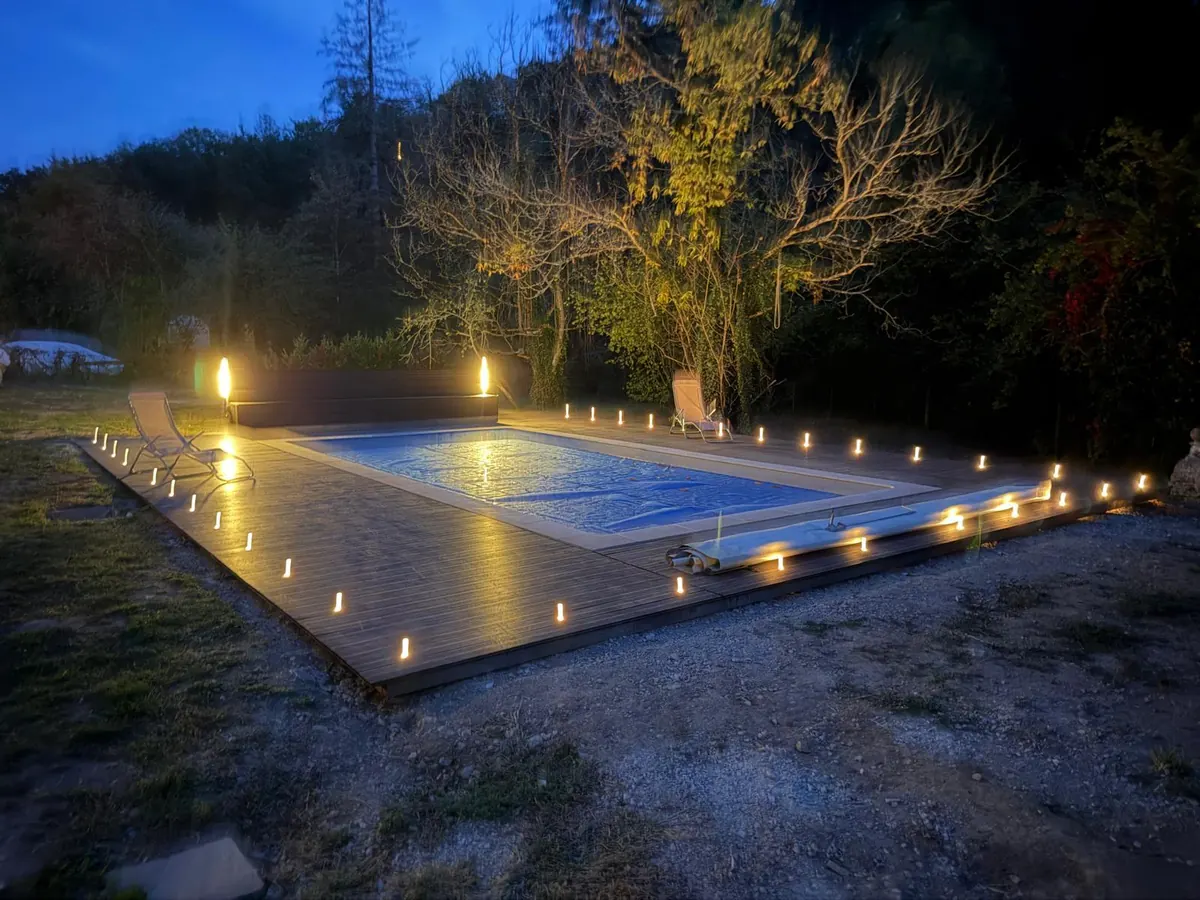 The pool at dusk with its cover on, the decking edged with rows of small glowing lights and two tall lamps at the far end, under trees lit gold