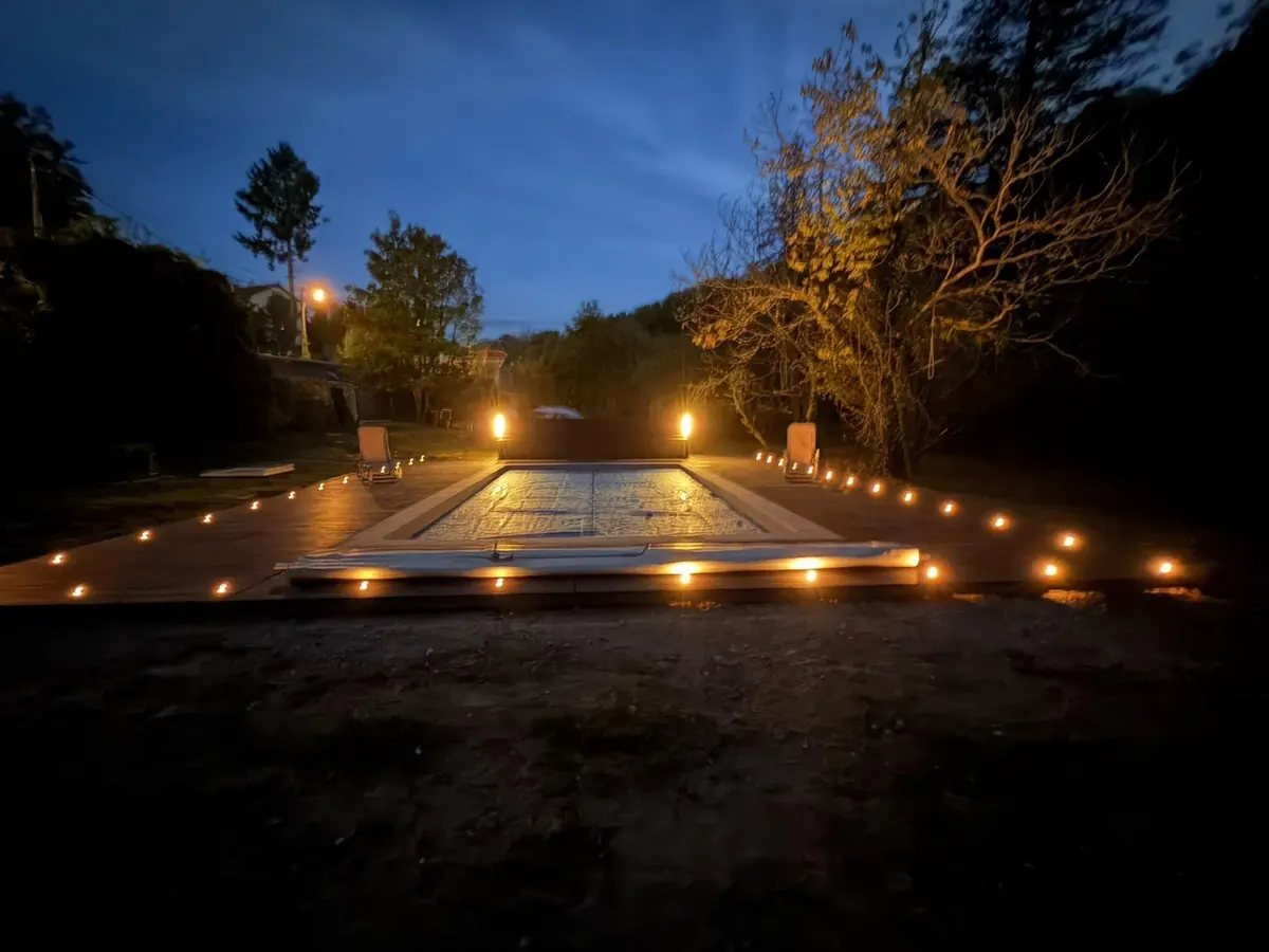 The pool from the end of the garden after dark, the decking outlined in warm lights and two tall lamps glowing either side of the far end